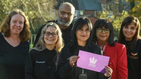 Six people are standing together looking at the camera and trees and bushes are behind them.