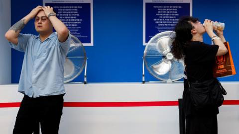 a man in a blue shirt with his head in hands and a woman wearing black drinking water, both standing in front of big fans.