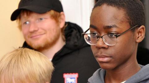 Ed Sheeran indoors wearing a black cap and black Ipswich Town jacket standing behind two boys