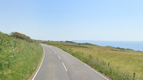 Google Street View of the coast road in summer. The two-way road is flanked by open meadows. To the right of the picture is the sea with a hazy Chesil Beach and Portland stretching out in the distance.