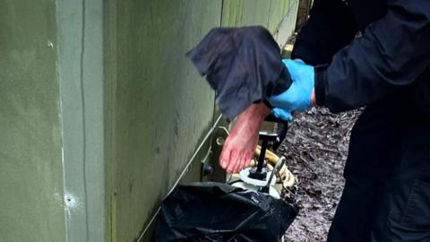 A police officer in dark blue overalls and wearing light blue latex gloves holds a replica of a human foot up to the camera.