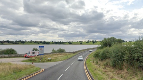 A road with a curved bend. On its left is a small car park with an ice cream van parked up. Beyond that is a large reservoir with green fields and trees around it.