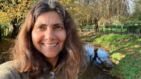 Dr Pallavi Devulapalli, former Green Party health spokesperson, standing outdoors near a small stream with autumn trees and a clear blue sky in the background.