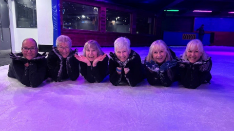 Six women lying on the ice, striking a pose by putting their hands to their faces. 