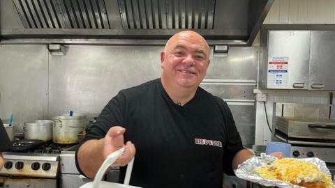 Mr Gribbin is standing in the cafe kitchen holding a chicken parmo. He is bald and wears a black t-shirt.