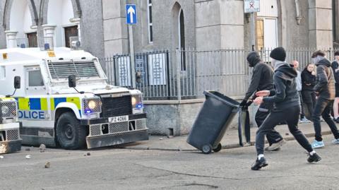 A number of masked individuals face large police vehicles on a street in the day time. One is pushing a wheelie bin. 