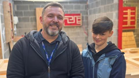 An older man smiles at the camera wearing a navy fleece and shirt, and a blue lanyard which says 'staff' on it. Standing next to him is a young girl, with short brown hair, wearing a blue coat, smiling proudly at the camera. Behind them is a construction classroom.