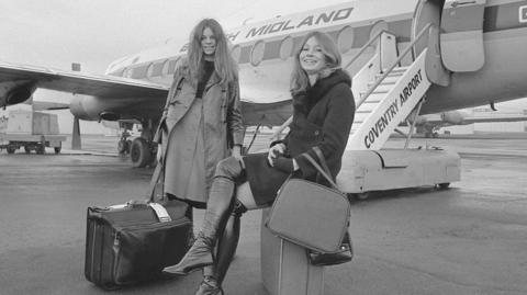 Two woman pose in front of a plane with suitcases in a black-and-white image. One with long fair hair is sitting on her suitcase and wearing tight black boots which come over the knee and a coat with a fur-trimmed collar.l The othe rhas long brown hair and wears a trench coat, a buttoned up skirt and a black top.