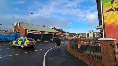 A police car in the middle of the road which has been closed with tape. A set of shops can be seen on the other side of the road.