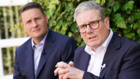 Starmer gestures with right hand while speaking in front of two microphones