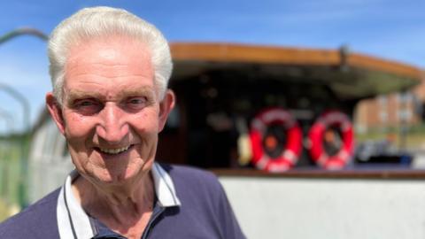 A smiling Rodney Bowles stands at the rear of the Boston Belle pleasure cruiser. He has swept-back, white hair and wears a blue shirt with white collar. Two red lifebuoy rings can be seen on the boat