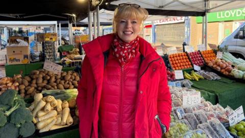 Lady smiling at camera wearing a bright pink coat and scarf, stood in front of a fruit and veg stand