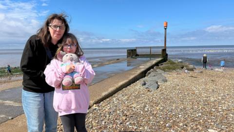 Colette and her daughter Amaya stand in front of the sea which is out in the picture exposing the mud flats. 