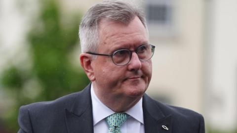 A man with short, cropped greying hair is seen in close up, with just his face and the tops of his shoulders visible. He is wearing dark-rimmed glasses, a dark blue suit jacket, white shirt and green patterned tie. 