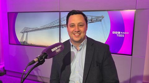 Ben Houchen sitting in the studio at BBC Tees. He is wearing a light blue shirt and a dark jacket, has short dark hair and is smiling. In front of him is a microphone with a purple cover and the BBC Tees branding in white letters. Behind him is a wide screen with the BBC Tees logo in white lettering on a purple background and an image of the Transporter Bridge