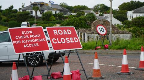 Traffic cones and three red and white signs block a road in a residential area. A number of white bungalows can be seen in the background. The rectangular signs read "SLOW CHECK POINT FOR AUTHORISED ACCESS" and "ROAD CLOSE". A circular "10" speed sign is also standing nearby. The signs are standing at an angle, as if they have been partially tipped over. 