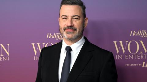 Jimmy Kimmel wearing a black suit with a white shit and a dark blue tie