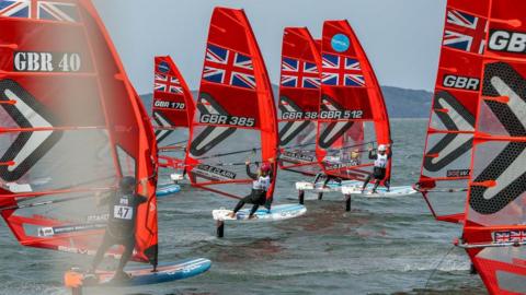 A fleet of Great Britain branded windsurfing boats they have bright red sails.