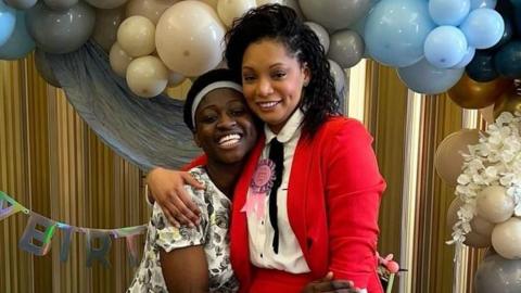 Two women stand in a warm embrace against a creamy yellow backdrop adorned with blue and white balloons. The woman on the left, who has dark hair, wears a white top with a black floral print. Beside her, the woman on the right wears a striking red suit over a white shirt.