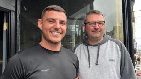 Andy Sheridan and Sam Morgan are both smiling at the camera. Andy wears a black round-neck t-shirt and has shore cropped hair. Sam is wearing a grey hoodie, has short dark hair and wears glasses.