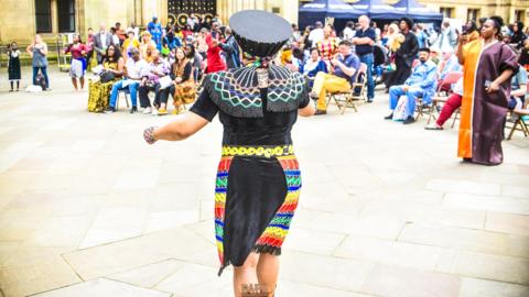A woman in colourful, traditional African clothing performs in front of a seated crowd in a city square. It's a bright and sunny day. The performer's back is to the camera.