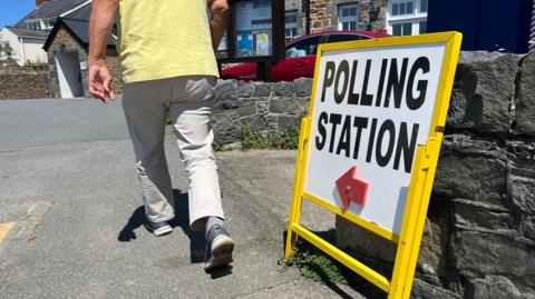 A man in a yellow top walks past a yellow polling station sign, with a red arrow in the direction he is walking.
