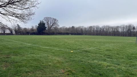 Grass sports field with obvious muddy spots. A single yellow cone marks out the corner of the pitch in the foreground. The far side of the field is bordered by large, mostly leafless trees.
