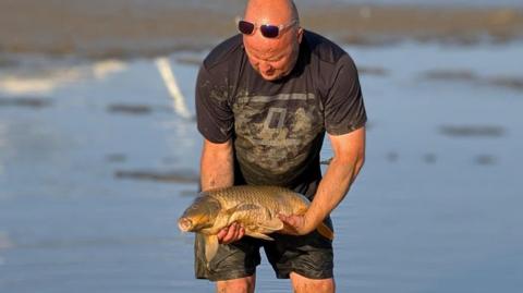 A man is standing in the middle of the lake, slightly crouched and holding a fish with both hands. The man is bald and has a pair of clear, square framed sunglasses on top of his head. He is wearing a black short sleeved t shirt which is covered in mud and a pair of black shorts, which are wet from walking through the water. He is holding a large carp, a brown and beige coloured fish, which has its mouth open. The background is blurred but shows some water and patches of dry land. 