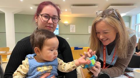 Gabriella Zubarevaite is looking straight at the camera with her infant son in her lap. Next to them is a staff member who is offering her son a toy car. Gabrielle is wearing a black jumper and has black glasses and red hair.