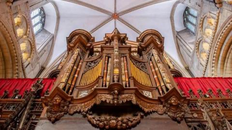 Gloucester Cathedral's organ pictured from below. It is wooden and has elaborate red and yellow designs on it, as well as wooden carved faces at the front of it. There are red curtains either side of the organ.