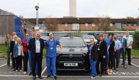 Plymouth hospital staff stand in front of the new X-Ray car. The car is black. Members of staff are stood either side of the car wearing their work uniform.