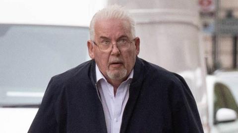 Andy Osborn arriving at court. He has short white hair and a goatee beard. He is wearing a dark zip up top and a light shirt underneath.