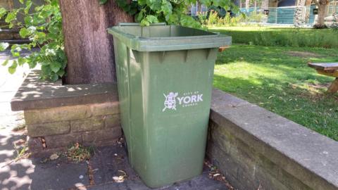 A green outdoor waste bin positioned beside a tree and a stone wall in a grassy park area. The bin is marked with the words “CITY OF YORK COUNCIL”.