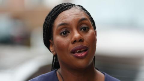 A head and shoulders shot of Kemi Badenoch speaking to someone off camera, looking straight ahead of her. She is wearing a dark purple top.