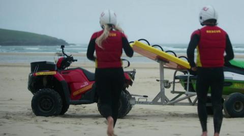 A picture of two people running towards a vehicle parked on the beach. There is two lifeguards pictured both wearing a black and red outfit.