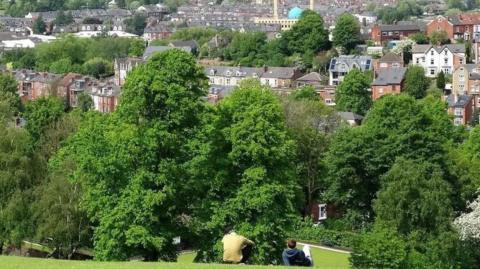 A view of Sheffield taken from the hill on Meresbrook Park. There are two people sitting on the hill and there are lots of trees in view. 