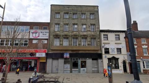 A Google street view image of some high street buildings with an old, dark brown four storey building in the centre of the frame. A "for sale" sign has been placed at the front. People can be seen walking past or sitting on public benches in the foreground.