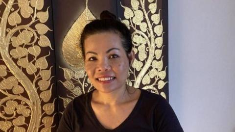 A woman in a black t-shirt with her hair in a bun standing in front of a decorative wall of golden leaves