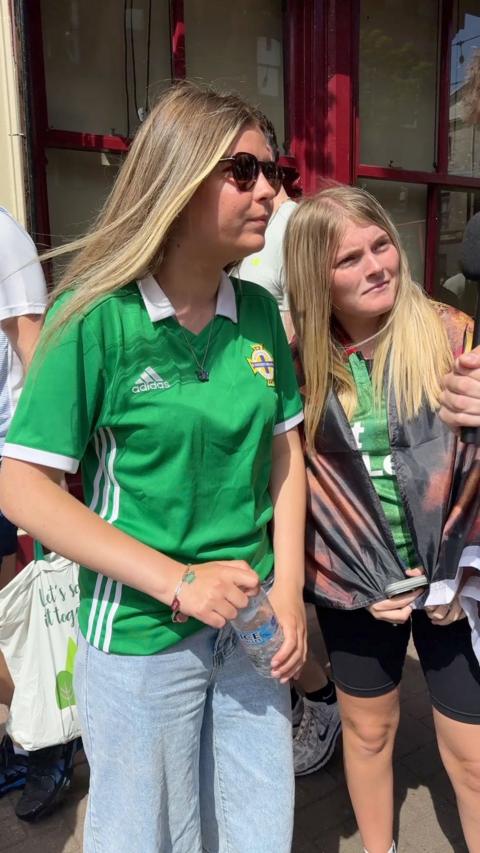 Two young girls with blonde hair wearing NI Football club jerseys stand at the side of the road looking at the interview who is off camera.