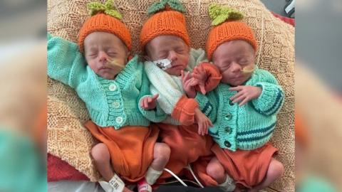 The triplets lying next to each other in green cardigans, orange Babygro's  and orange woollen hats with green knitted leaf tops.