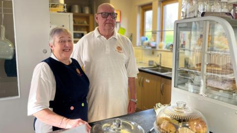 Anne Kemp smiling at the camera next to her husband Derek Kemp behind the counter at Bede's Bakehouse. They are wearing white t-shirts with the red and yellow logo of St Peter's Church. Anne has short grey hair and is wearing a dark blue apron. Derek has black-rimmed glasses. There is a range of cakes and bakes in front of them.