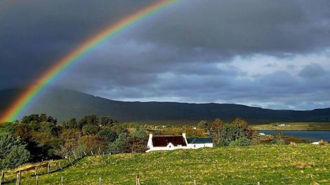 Rainbow amid a grey sky above a white house and field in the countryside with mountains in the distance