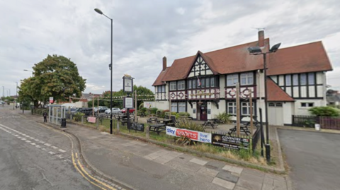 A pub with a red roof with dark beams near the side of a road. A bus stop is in front of the pub.