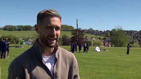 Woakes with brown hair and a brown top is on the left of the photo. A field and other people are in the background, while trees and rooftops are in the distance. 