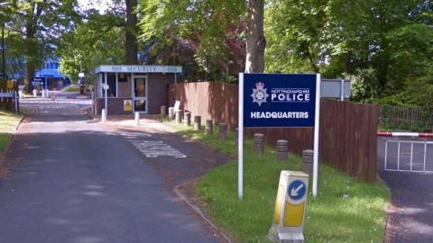 A street view of the entrance to Nottinghamshire Police HQ showing a sign that reads Nottinghamshire Police Headquarters with. A security box and gate can be seen in the background, leading to a main building. 