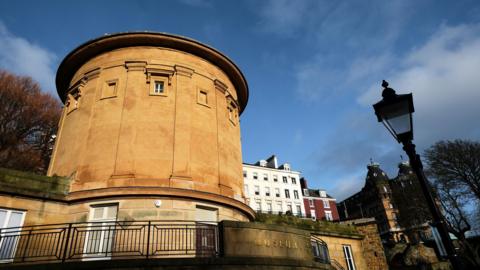 A circular sandstone building with a walkway beneath encased by a black railing. On the railing stands a brick wall with the word 'museum' attached in bold gold letters.