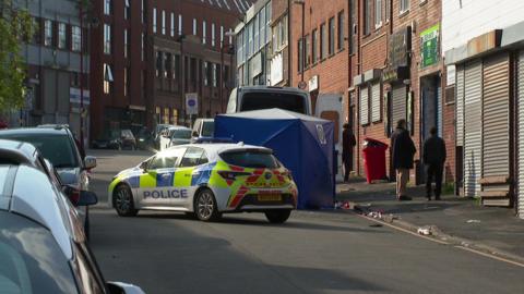 A street with cars parked on the left. In the middle of the road is a police car, which is next to a blue tent. There is a white van behind it with its back doors open. To the right is a pavement with litter strewn on the floor and there are three people standing on the pavement with their backs to the camera. 