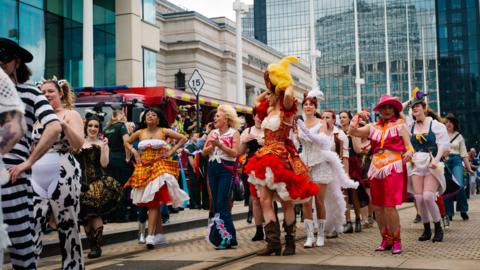 A group of people in a parade dressed in various outfits - mostly colourful costumes - as they walk through a city centre. 