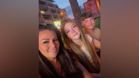Three people sit close together outdoors in the evening, facing the camera for a selfie. A tree trunk is directly behind them, and a multi-story building with balconies and lit windows appears in the background. Streetlights and signage glow softly as the sky turns dusky blue. The three individuals are positioned shoulder-to-shoulder, with one slightly in front holding the camera, and the other two seated beside them.