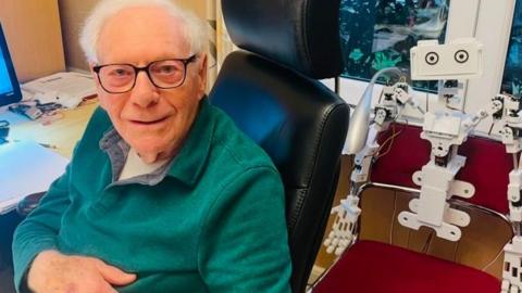 Malcolm Stern is seated in the office chair and is looking toward the camera.
He is wearing dark-framed glasses and a green quarter-zip fleece or sweater over a light-colored shirt.